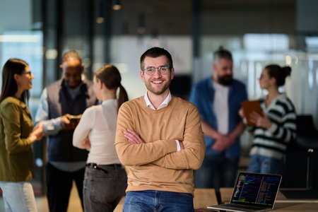 Portrait of a confident young businessman wearing glasses and a beige sweater, standing with arms crossed in a modern office while colleagues collaborate in the background.の写真素材