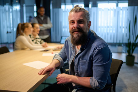 Portrait of a smiling confident businessman with beard sitting at conference table during a team meeting in a modern office, symbolizing leadership, collaboration, and teamworkの写真素材