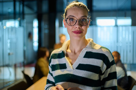Portrait of a confident businesswoman wearing glasses and striped sweater standing in a modern office environment, representing leadership, intelligence, focus, and success in the workplace.の写真素材