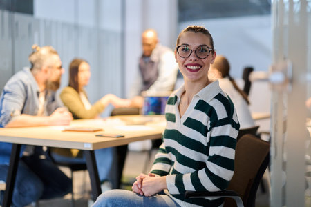 Smiling businesswoman sitting confidently in a modern office while her team collaborates in the...
