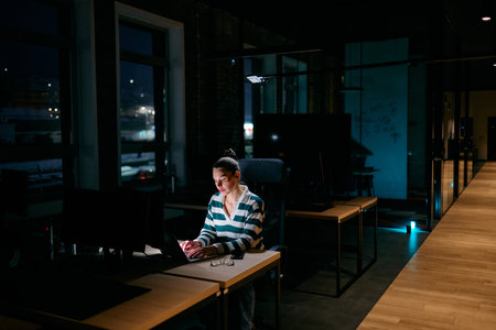 Concentrated businesswoman working late at night on laptop in dimly lit office, representing dedication, focus, and modern remote work culture under low light conditions.の写真素材