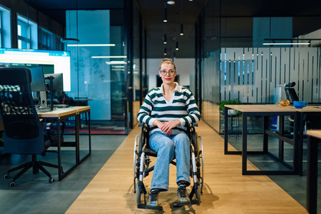 A confident smiling business woman in a wheelchair sitting in a bright modern office hallway,...