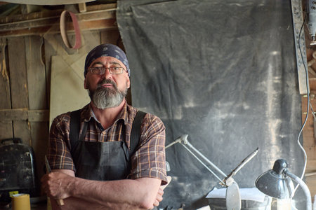 Portrait of a craftsman in his workshop, wearing glasses and a bandana, with a beard, looking at the camera, indoor scene, natural lighting, skilled worker.の写真素材