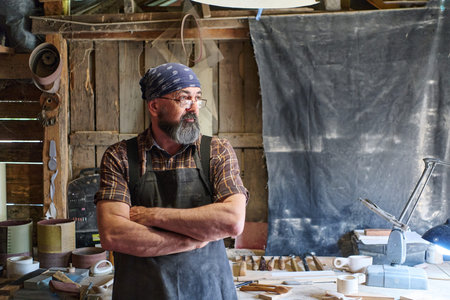 Artisan woodworker in his workshop, wearing a bandana and apron, arms crossed, looking thoughtfully,...