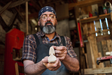 Woodcarver carefully shaping a small wooden figure in his workshop, artisan crafting a wooden sculpture, hobbyist working with hand tools, creating art in a rustic studio environment.の写真素材
