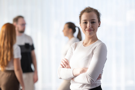 Portrait of a confident woman standing with arms crossed and smiling at the camera while her colleagues interact in the blurred background.の写真素材