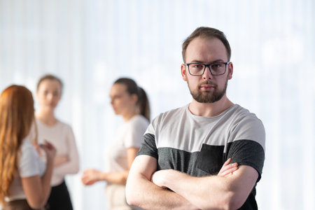 Portrait of a confident young man with glasses smiling and standing with folded arms while his colleagues talk in the background. Concept of leadership, teamwork, and success in a modern professional environment.の写真素材