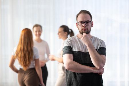 Portrait of a confident young man wearing glasses and smiling while standing with his colleagues in the background during an informal business meeting.の写真素材