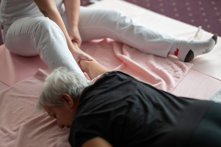 A physiotherapist assisting an older woman with a shoulder and arm stretch while lying on a mat. Rehabilitation and physiotherapy session concept focusing on mobility, flexibility, pain relief, and holistic wellness care.の写真素材