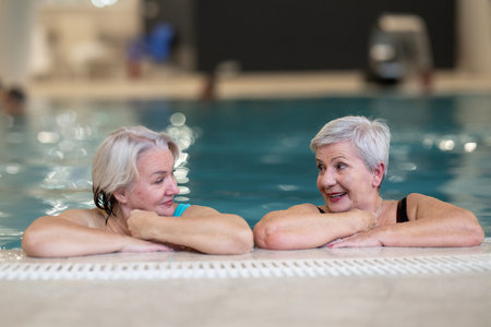 Two smiling senior women enjoying leisure time in an indoor swimming pool, leaning on the poolside. Concept of friendship, wellness, active lifestyle, relaxation, and healthy aging.の写真素材
