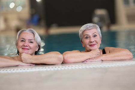 Two smiling senior women enjoying leisure time in an indoor swimming pool, leaning on the poolside. Concept of friendship, wellness, active lifestyle, relaxation, and healthy aging.の写真素材