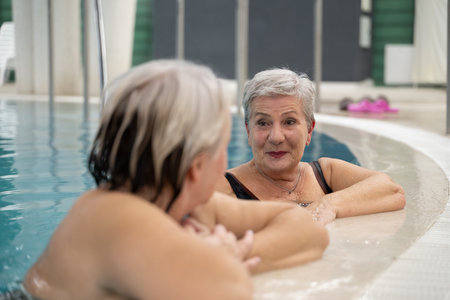 Two smiling senior women enjoying leisure time in an indoor swimming pool, leaning on the poolside. Concept of friendship, wellness, active lifestyle, relaxation, and healthy aging.の写真素材