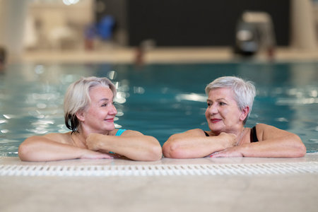 Two smiling senior women enjoying leisure time in an indoor swimming pool, leaning on the poolside. Concept of friendship, wellness, active lifestyle, relaxation, and healthy aging.の写真素材