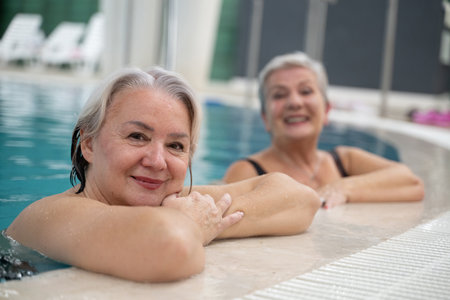 Two smiling senior women enjoying leisure time in an indoor swimming pool, leaning on the poolside. Concept of friendship, wellness, active lifestyle, relaxation, and healthy aging.の写真素材