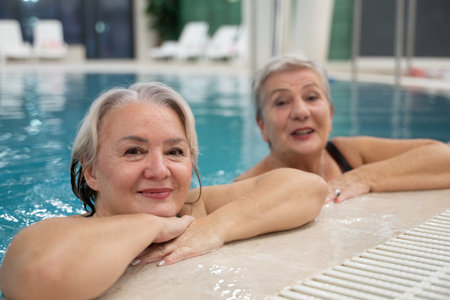 Two smiling senior women enjoying leisure time in an indoor swimming pool, leaning on the poolside. Concept of friendship, wellness, active lifestyle, relaxation, and healthy aging.の写真素材