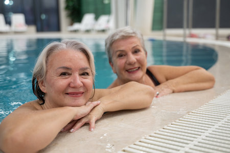 Two smiling senior women enjoying leisure time in an indoor swimming pool, leaning on the poolside. Concept of friendship, wellness, active lifestyle, relaxation, and healthy aging.の写真素材
