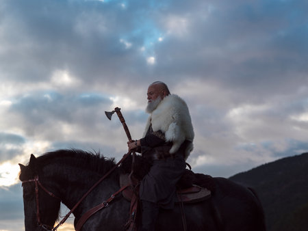 A fierce Viking warrior on horseback, wielding an axe, stands against a dramatic sunset sky with clouds and mountains, embodying strength, exploration, and ancient legend.の写真素材