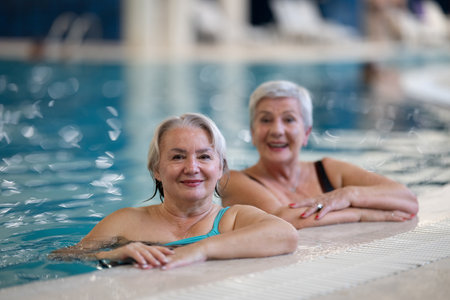 Two smiling senior women enjoying leisure time in an indoor swimming pool, leaning on the poolside. Concept of friendship, wellness, active lifestyle, relaxation, and healthy aging.の写真素材