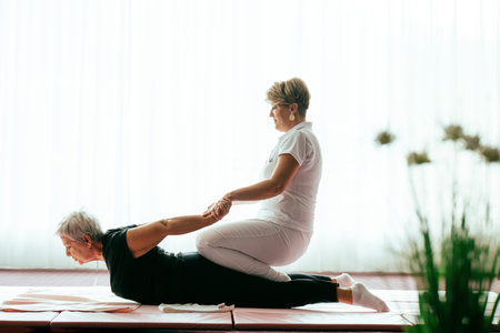 Professional female physiotherapist supporting an older woman during a therapeutic stretching exercise on a mat. Rehabilitation and physiotherapy concept focused on mobility, flexibility, recovery, and holistic health care for seniors.の写真素材