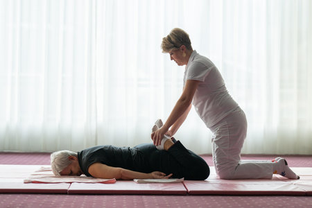 Professional female physiotherapist helping an older woman stretch her legs while lying face down on a mat. Rehabilitation and physiotherapy concept focusing on mobility recovery, flexibility, and holistic wellness care for seniors.の写真素材