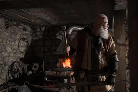 A Viking blacksmith with a white beard and braided hair works intently at a forge, hammering glowing metal on an anvil, with flames flickering in the background of a stone workshop.の写真素材