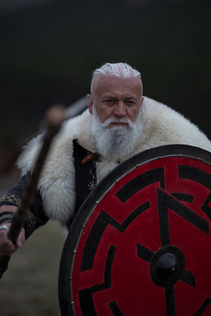 Viking warrior with axe standing on mountain cliff, wearing fur cloak and leather armor, overlooking dramatic landscape. Historical Nordic fighterの写真素材