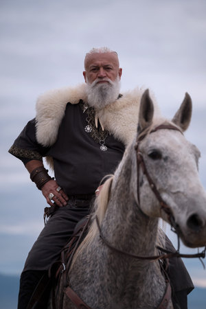 A majestic Viking chieftain on a dappled grey horse, clad in a fur cloak, surveys a desolate,...