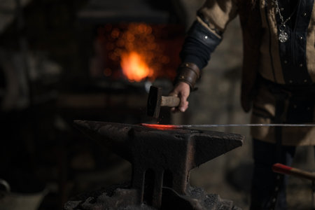 A Viking blacksmith with a white beard and braided hair works intently at a forge, hammering glowing metal on an anvil, with flames flickering in the background of a stone workshop.の写真素材
