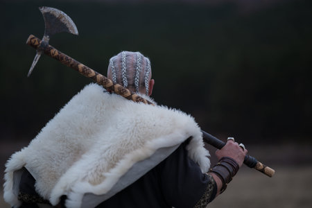 Viking warrior with axe standing on mountain cliff, wearing fur cloak and leather armor, overlooking...