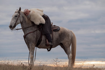 Elderly Viking warrior man with a white beard, embracing a gray horse warmly, dressed in traditional attire against a tranquil sky, depicting companionship and serenityの写真素材