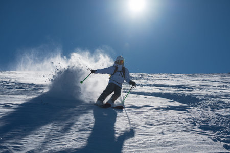 Skier skiing off piste through fresh powder on a sunny mountain slope, kicking up snow as she...