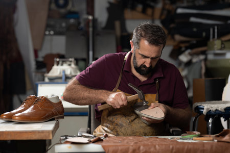 Leather craftsman forging shoes in a busy workshop with hammer, awl, and leatherの写真素材