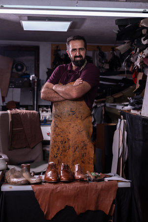 Leather craftsman stands with arms crossed in his workshop, displaying handmade shoes