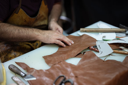 Leather craftsman at work shaping brown leather pieces with tools and patterns on a busy worktable