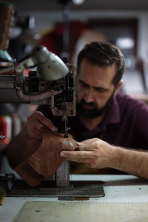 Leather craftsman sewing leather piece on an industrial machine in a busy workshop