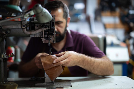 Leather craftsman sewing leather piece on a vintage industrial sewing machine in workshop, at the...