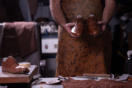 Craftsman holds polished brown leather shoes in a busy workshop, surrounded by tools and leather scrapsの写真素材