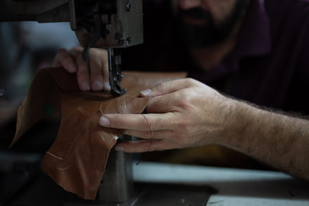 Craftsman guides brown leather under a sewing machine, stitching a piece with careful, precise...