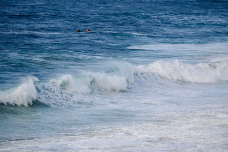 Powerful ocean waves roll toward shore as distant surfers ride swells under a bright blue skyの写真素材