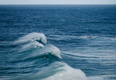 Powerful ocean waves crash as surfers ride towering swells under a clear blue sky