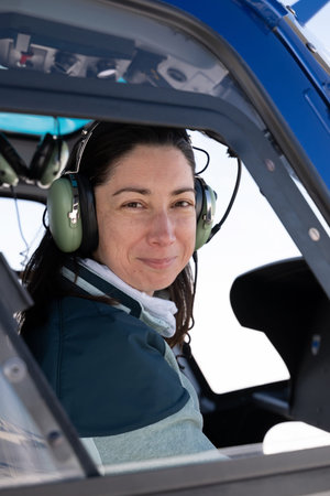 Female helicopter pilot wearing headset sits in cockpit, smiling in blue helicopterの写真素材