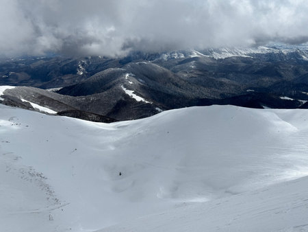 Snow-covered mountain ridge overlooking forested valleys and moody winter clouds in a remote alpine...