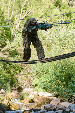 Camouflaged Sniper Soldier Crossing Suspended Rope Bridge Over Stream During Woodland Reconnaissanceの写真素材