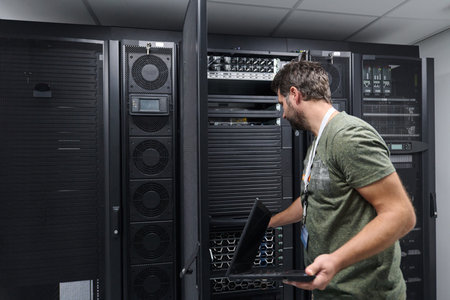 Male IT technician works on servers in a data center server room with a laptop performing...