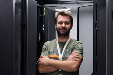Confident IT technician standing in server room with arms crossed among data racks and equipment