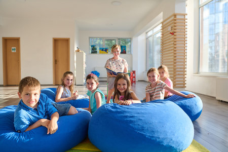 Children and teacher reading together on blue bean bags in a bright classroom loungeの写真素材