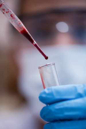 Young medical technician dropping blood sample into test tube in clinical laboratory with gloved hands