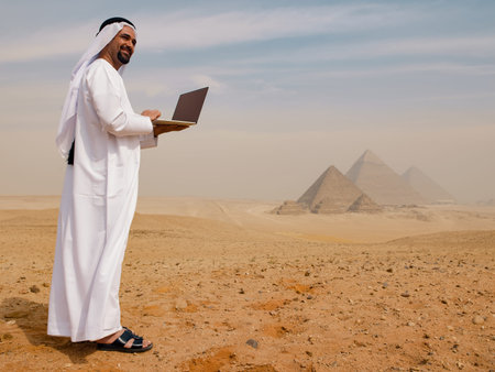 Smiling Arab businessman in traditional white thobe using laptop in Egyptian desert with Giza pyramids behind him ideal for Middle East technology and