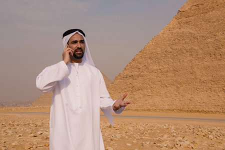 Animated Arab businessman in traditional white thobe gesturing during a phone call on his smartphone before the Giza pyramids in Egypt ideal for