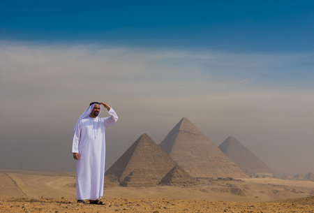 Proud arab man wearing traditional white kandura shielding eyes and gazing toward the iconic Giza pyramids across vast Egyptian desert ideal for